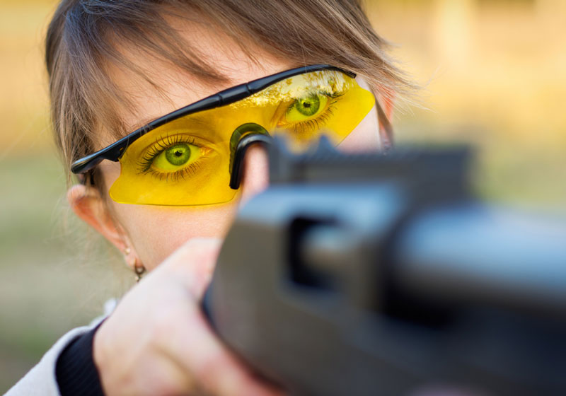 A young girl with a gun for trap shooting and shooting glasses aiming at a target