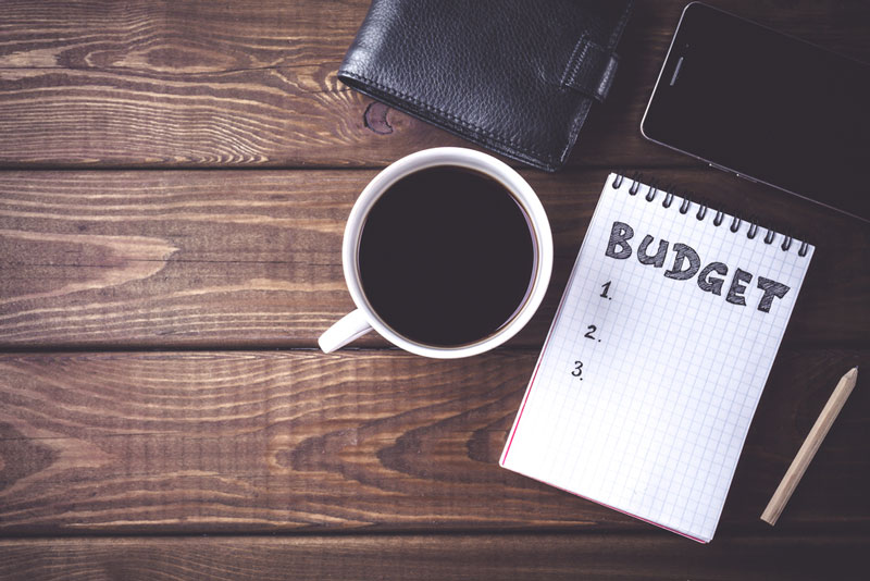 Top view of notepad with word Budget, mobile phone, cup of coffee, pouch on wooden background