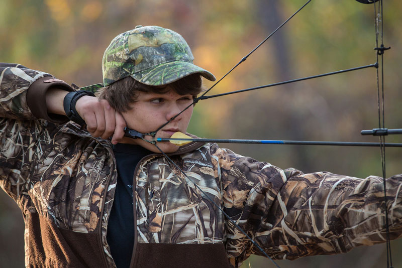 Young boy in camo taking aim with bow and arrow